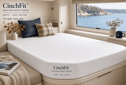 A fitted sheet by CinchFit shown on a mattress in a real bedroom setting with a window view of water and rocks, pillows, and decor. The sheet is white and branded with CinchFit text near the lower front corner.