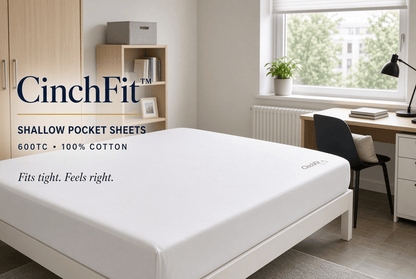 A white fitted sheet on a mattress in a bright bedroom setting, with text overlay reading 'CinchFit' and 'Shallow Pocket Sheets 600TC • 100% Cotton'. The room includes a window, desk, chair, and shelving.