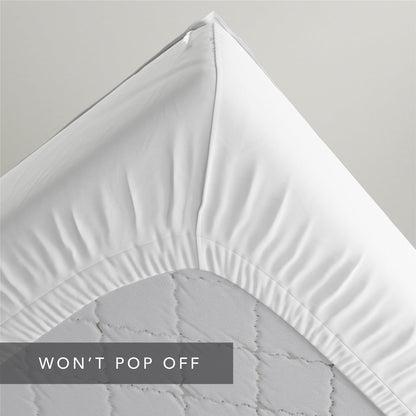 A close-up of a white  Quickzip Fitted Sheet corner pulled tightly over the corner of a light gray quilted mattress, with a dark gray banner at the bottom reading 'WON'T POP OFF'.
