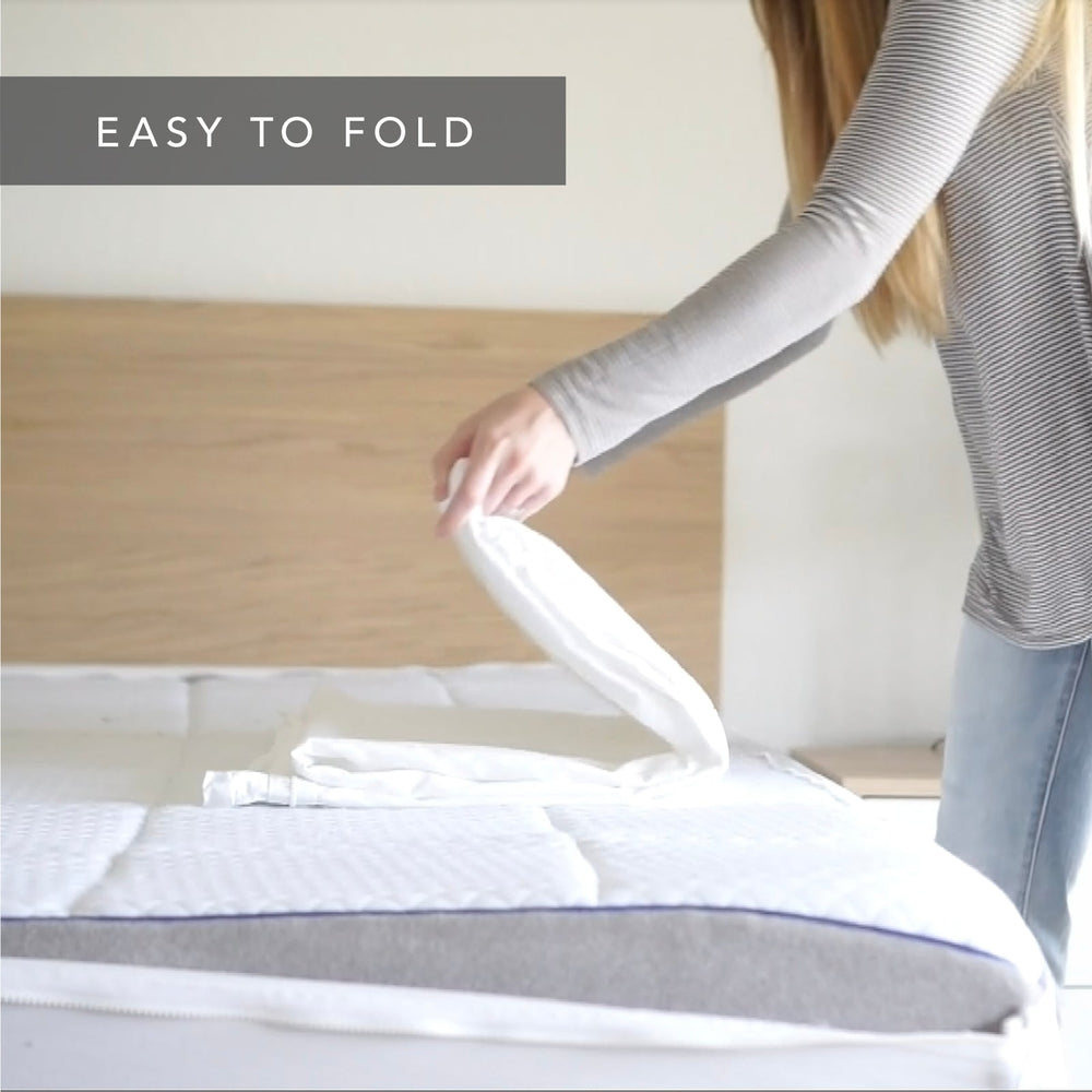 A woman in a gray striped long-sleeve shirt and blue jeans is folding a white  Quickzip Fitted Sheet on a mattress. The text 'EASY TO FOLD' is displayed in a gray banner at the top left.