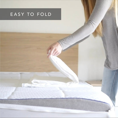 A woman in a gray striped long-sleeve shirt and blue jeans is folding a white  Quickzip Fitted Sheet on a mattress. The text 'EASY TO FOLD' is displayed in a gray banner at the top left.