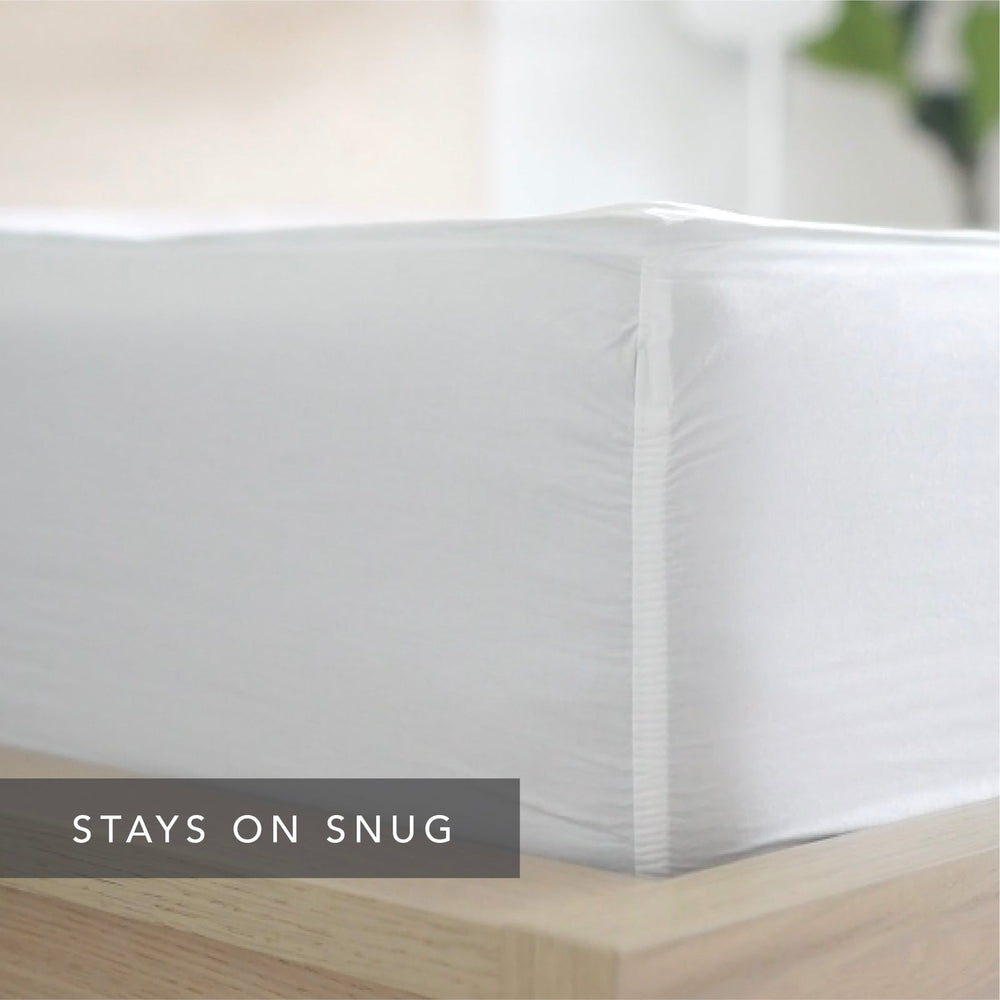 A close-up of a white  Quickzip Fitted Sheet snugly covering the corner of a mattress on a light wood bed frame, with the text 'STAYS ON SNUG' overlaid at the bottom left.