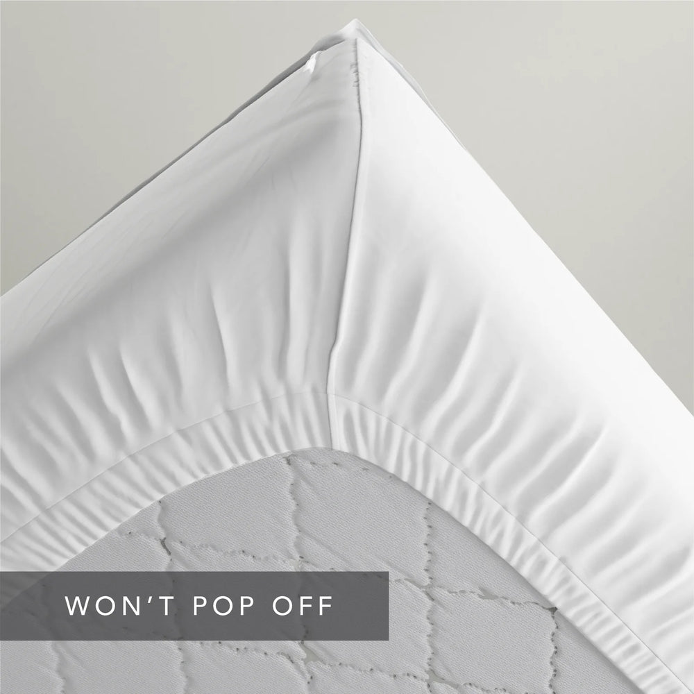 A close-up of a white fitted sheet draped over the corner of a mattress, showing the gathered elasticized edge and quilted mattress surface. A dark gray text banner at the bottom reads 'WON'T POP OFF'.