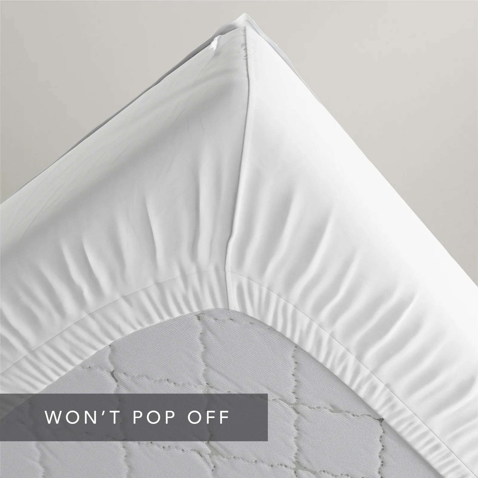 A close-up of a white fitted sheet draped over the corner of a mattress, showing the gathered elasticized edge and quilted mattress surface. A dark gray text banner at the bottom reads 'WON'T POP OFF'.
