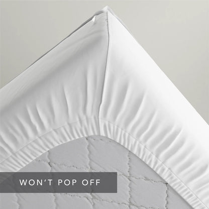 A close-up of a white fitted sheet draped over the corner of a mattress, showing the gathered elasticized edge and quilted mattress surface. A dark gray text banner at the bottom reads 'WON'T POP OFF'.