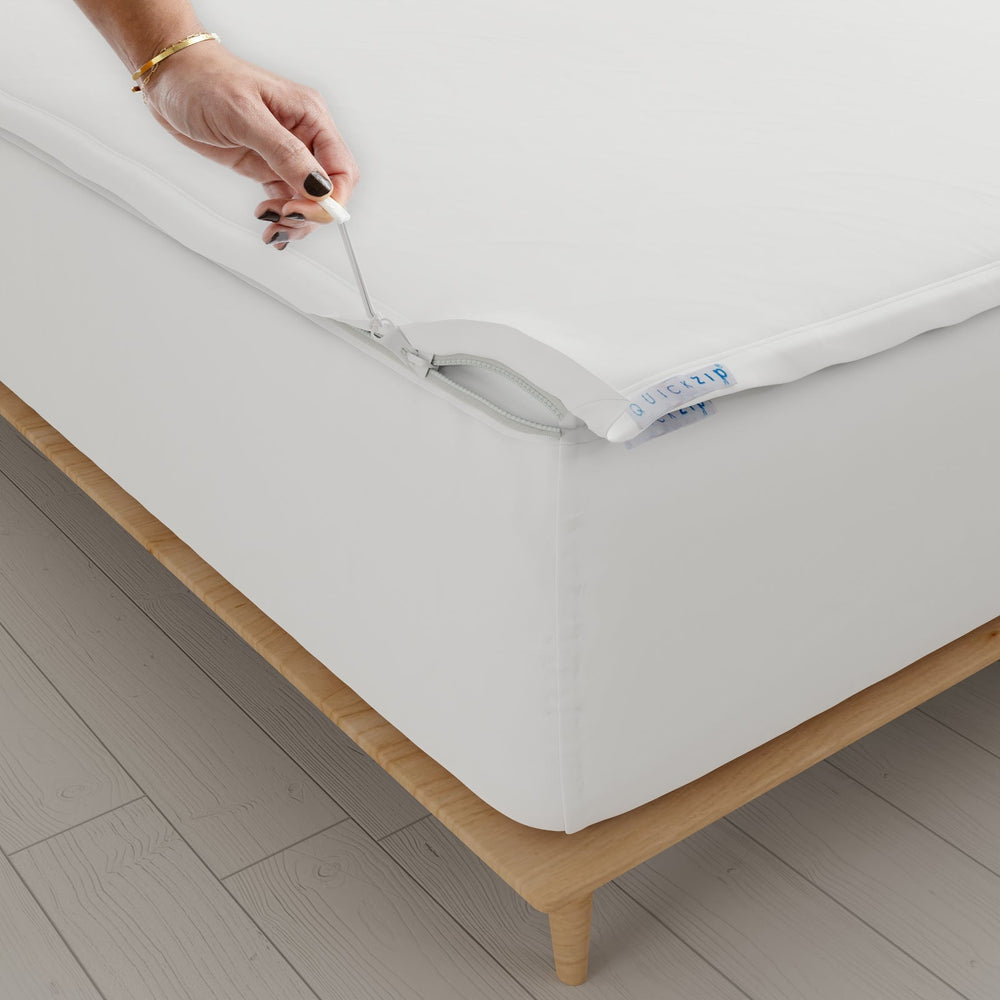 A close-up of a white  Quickzip Fitted Sheet being fitted over a mattress corner on a wooden bed frame, with a hand pulling a metal hook-and-loop fastener tab.